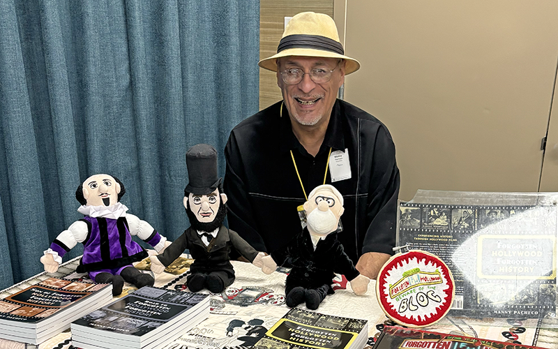 03-Story_01_LightsCameraAction A smiling man in a hat sits behind a table with books, three puppets, and a Hollywood-themed display at an event. A blue curtain and tan wall are in the background.