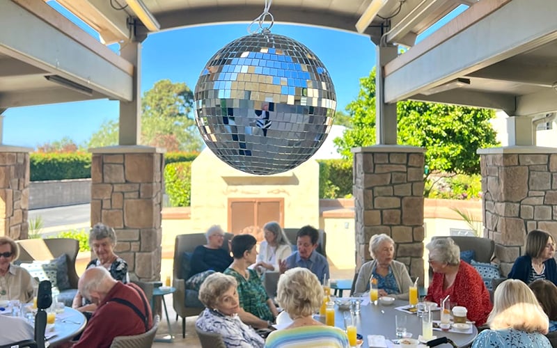 03-Story_02_SipSipHooray A group of seniors sit at tables under a patio with a disco ball hanging above them, enjoying drinks and conversation in a relaxed, sunny outdoor setting.