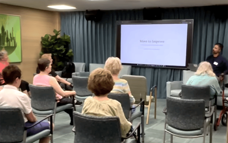 03-Story_03_Move Speech A group of older adults sit in a room facing a presenter and a large screen displaying Move to Improve. The room has chairs, a plant, a painting, and blue curtains.