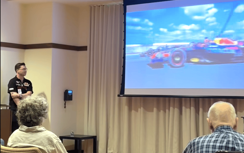 03-Story_03_NeedforSpeed A man stands by a podium in a conference room while a racing car is shown on a projector screen to an audience of seated people.