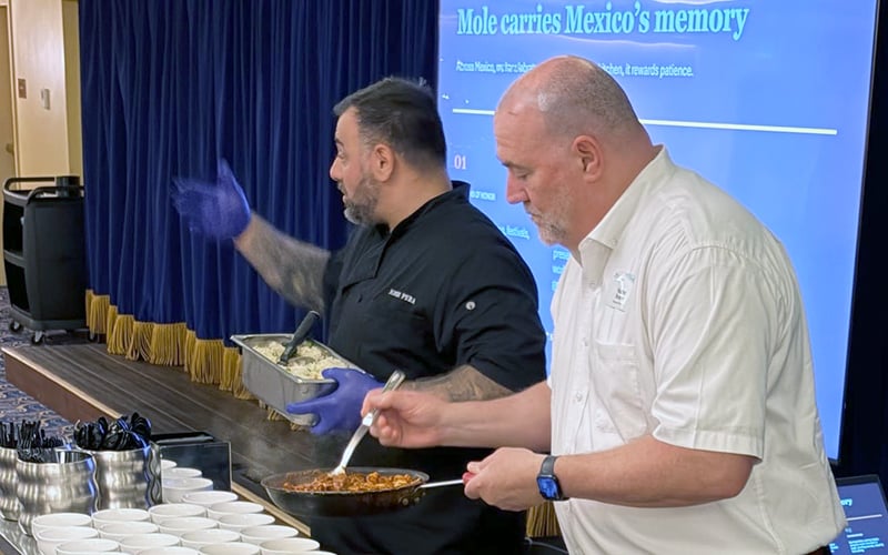 Two men serve food into cups at a table. One man, in a black chef coat and purple gloves, gestures while holding a tray. The other, in a white shirt, spoons food from a pan. A large blue screen is visible in the background.