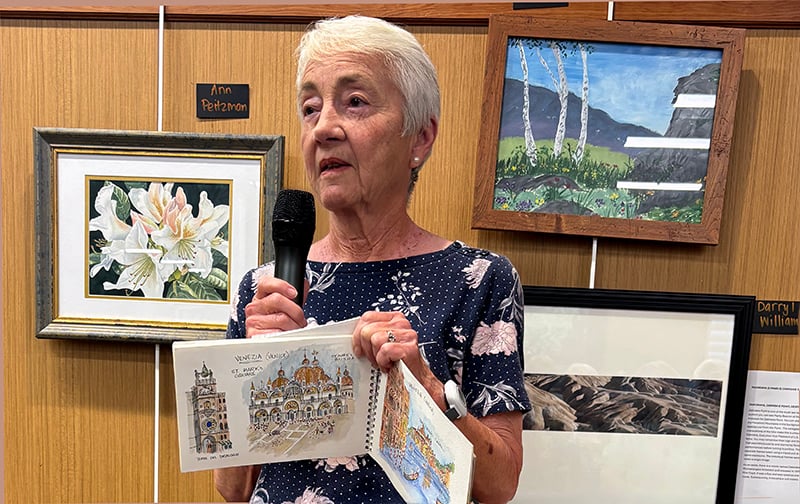 03-Story_04_Artists Gallery An older woman with short gray hair speaks into a microphone while showing a sketchbook with drawings. She stands in front of a display of framed artwork on a wooden wall.
