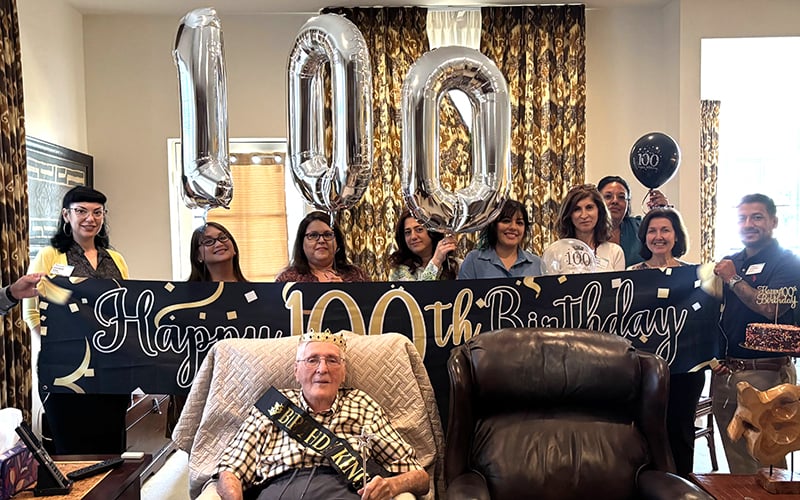 03-Story_05_CelebratedCentury A group celebrates a 100th birthday with balloons, a banner reading Happy 100th Birthday, and an elderly man in a sash and crown sitting in front of the group.