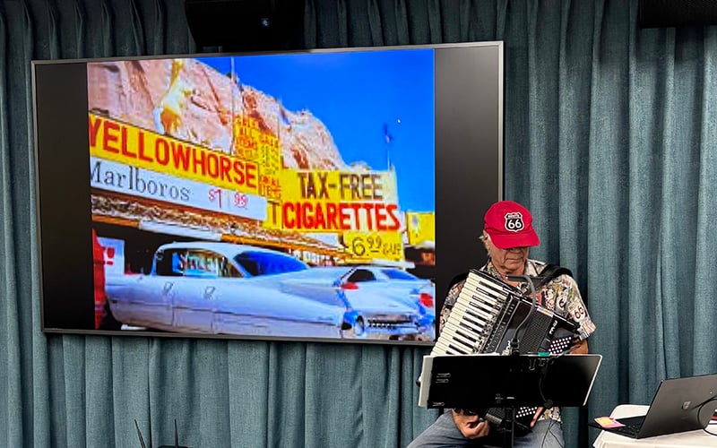 03-Story_05_Route66 Preso A man wearing a Route 66 cap plays an accordion in front of a screen displaying a vintage car parked at a store advertising Marlboros and tax-free cigarettes. Blue curtains are in the background.