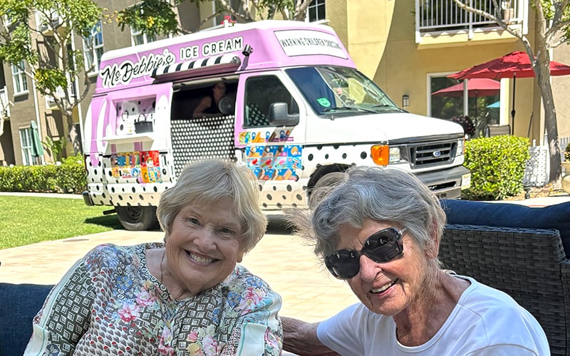 Two smiling elderly women sit outdoors in front of a pink and white ice cream truck on a sunny day, with green grass and apartment buildings in the background.