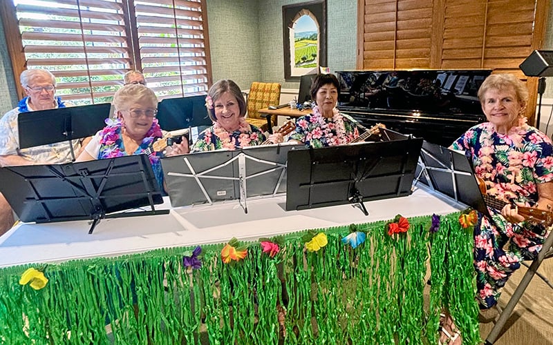 Five smiling seniors in floral shirts and leis play ukuleles behind music stands decorated with green grass and colorful flowers, sitting indoors near a piano and wooden blinds.