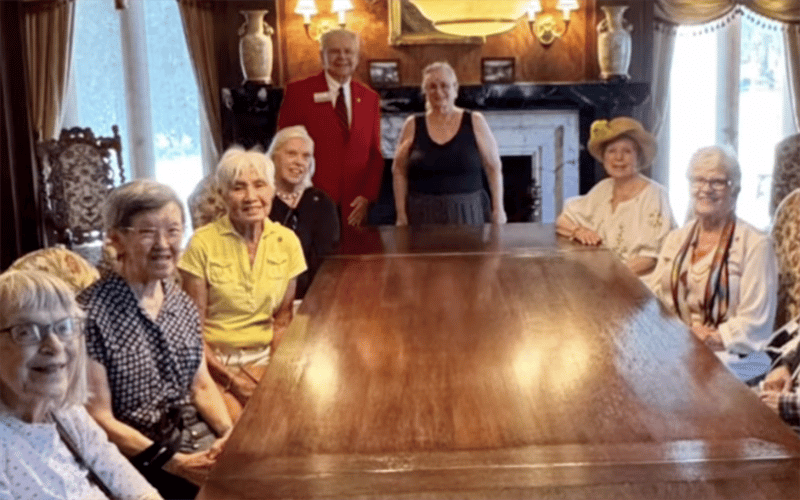 03-Story_06_TournRoses A group of older adults, mostly women, sit and stand around a large wooden table in an elegant, warmly lit room with ornate decor and a fireplace in the background.