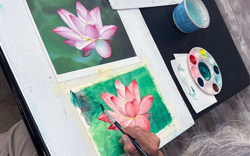 03-Story_06_WatercolorClass A person paints a pink lotus flower on green background, referencing a photo. Nearby are a paint palette, cup of water, and paper towels.