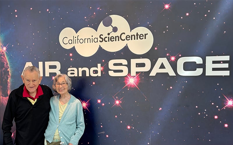 03-Story_07_CA Science Center An elderly man and woman stand smiling in front of a backdrop with California Science Center AIR and SPACE and a starry space design.