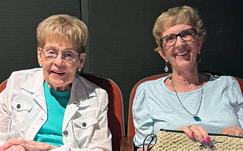Two older women with short hair and glasses sit side by side, smiling. One wears a white jacket over a teal top, and the other wears a light blue shirt, necklace, and holds a woven bag. They are seated against a dark background.