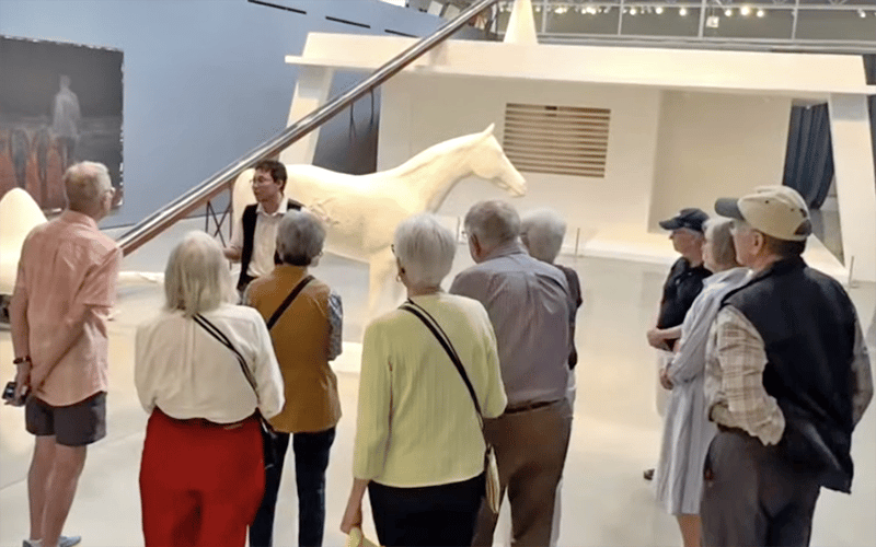 03-Story_07_Wende2 A group of older adults listens to a guide in a museum, standing near a large white horse sculpture and modern art displays.