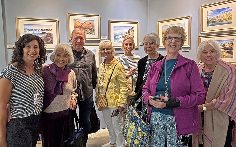 A group of eight smiling people pose together in front of a wall with framed landscape paintings in an art gallery.