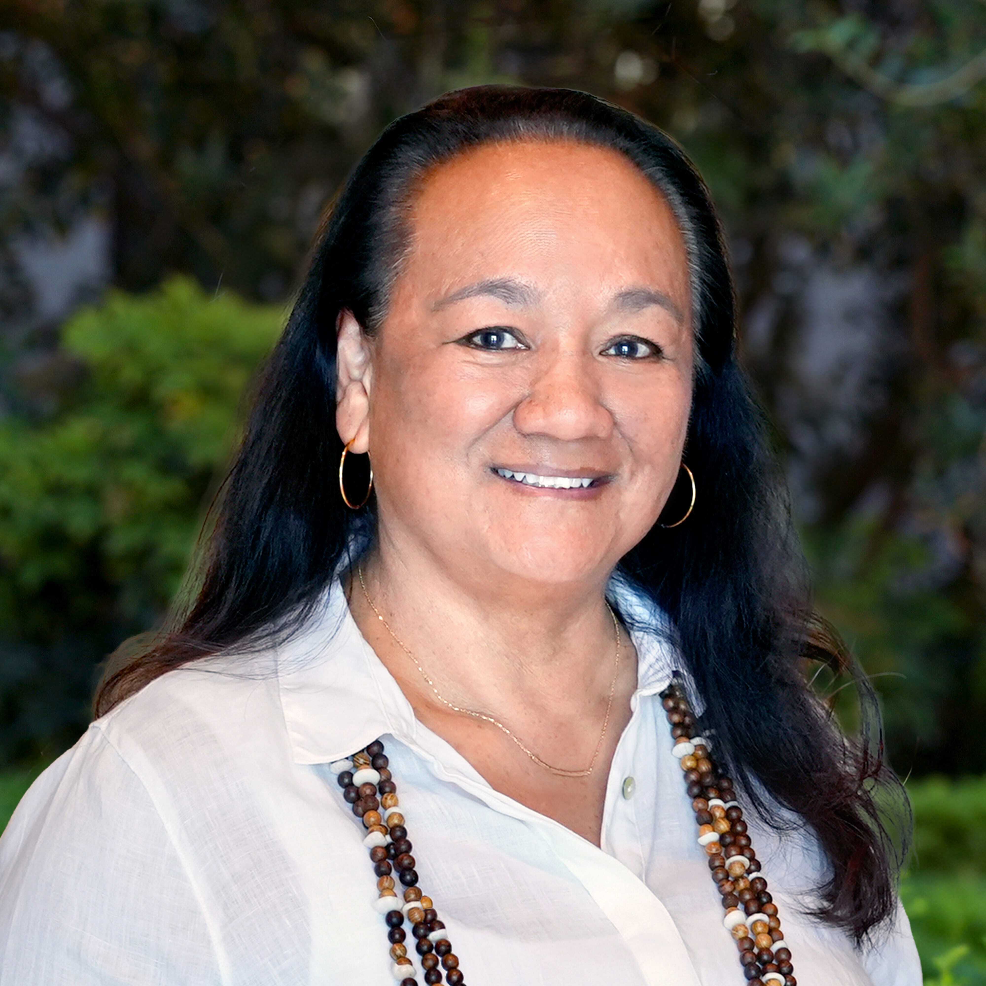 A woman with long dark hair, wearing a white shirt, gold hoop earrings, and a beaded necklace, smiles while standing outdoors with green foliage in the background.