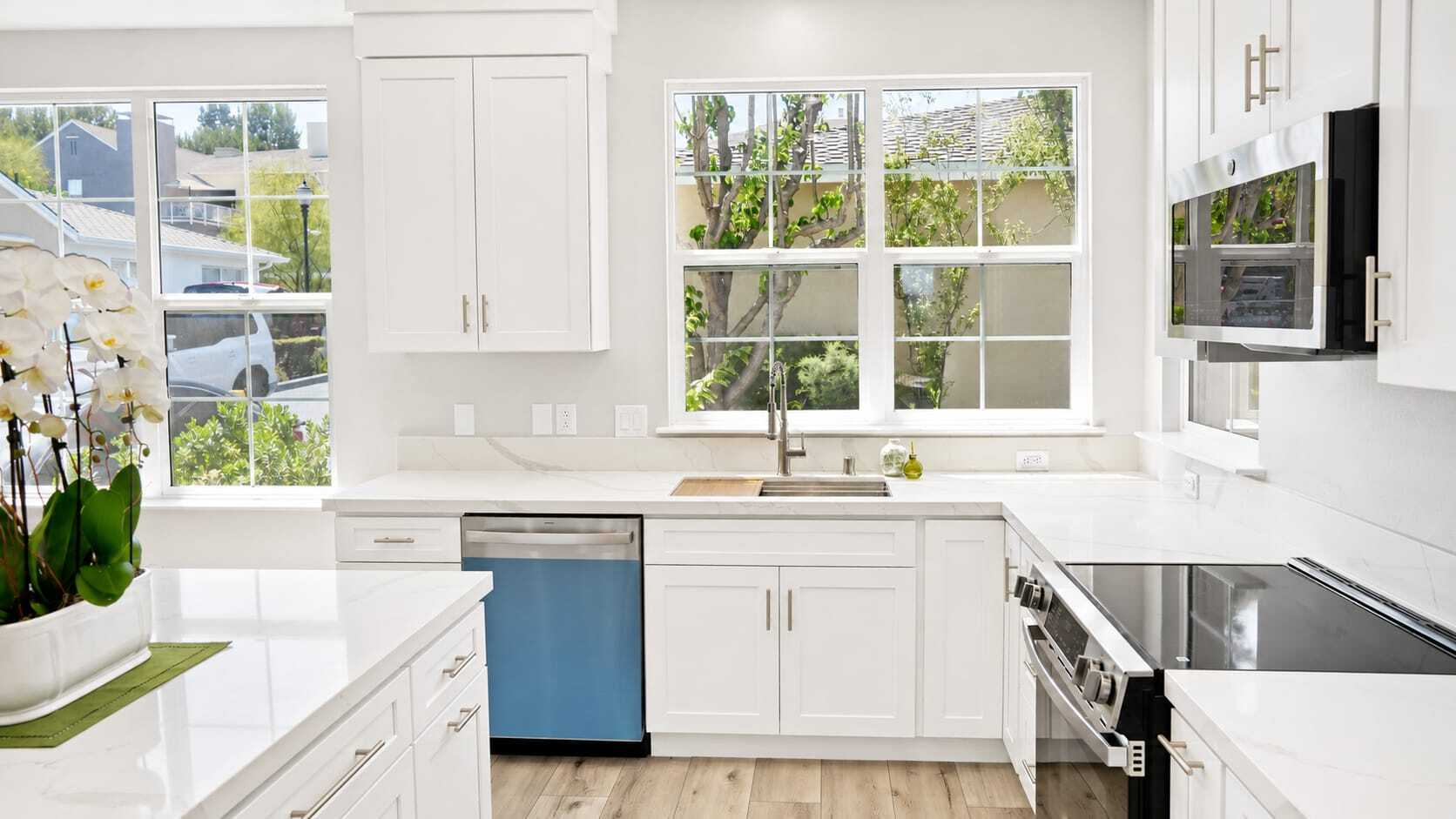 Bright modern kitchen with white cabinets, stainless steel appliances, a large window above the sink, light wood flooring, and a potted orchid on the island. Natural light fills the space.