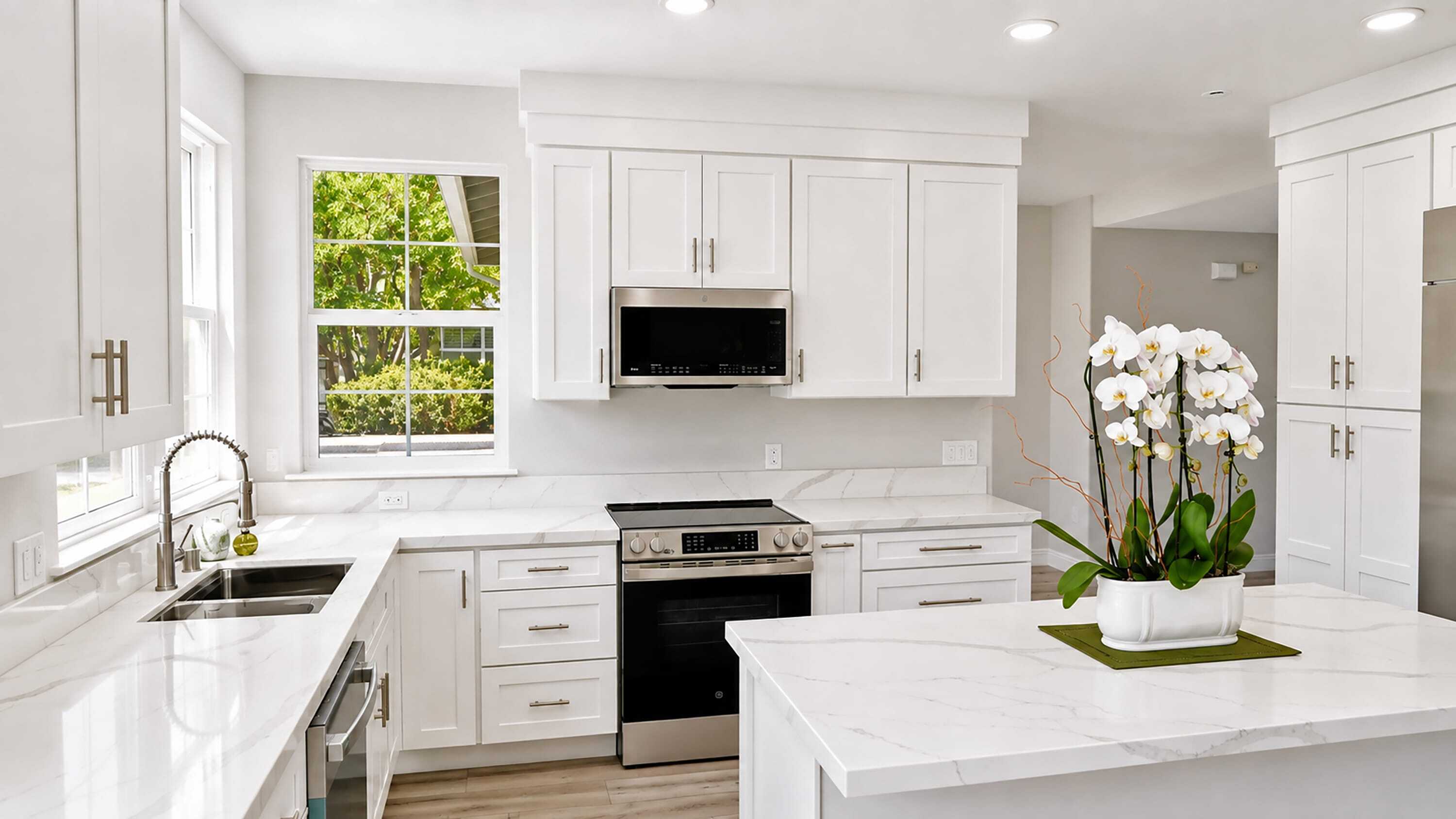 Bright modern kitchen with white cabinets, marble countertops, stainless steel appliances, a sink under a window, and a kitchen island with a potted white orchid on top.