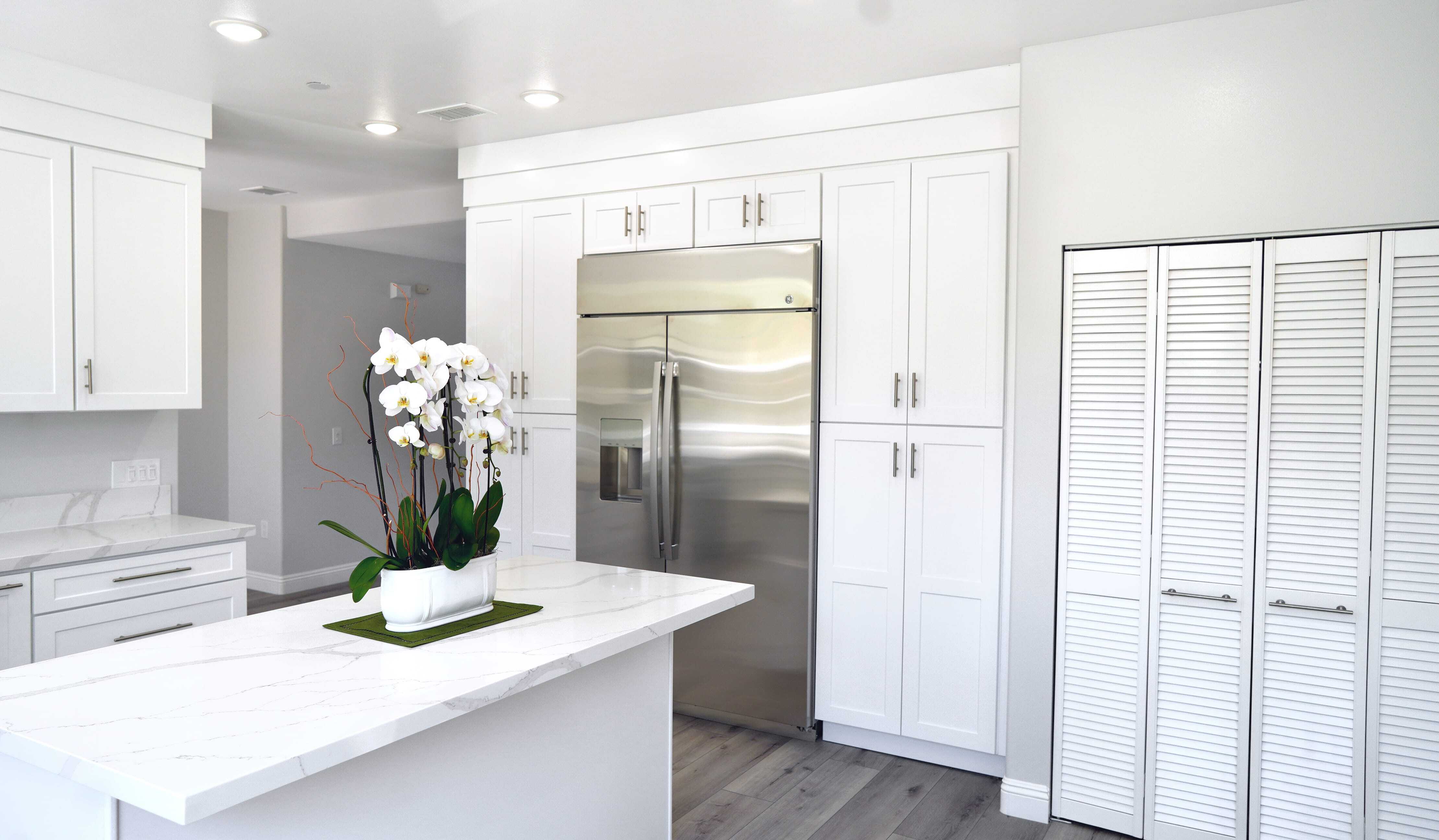Modern kitchen with white cabinets, stainless steel refrigerator, marble island with a potted orchid, light wood floors, and folding pantry doors.