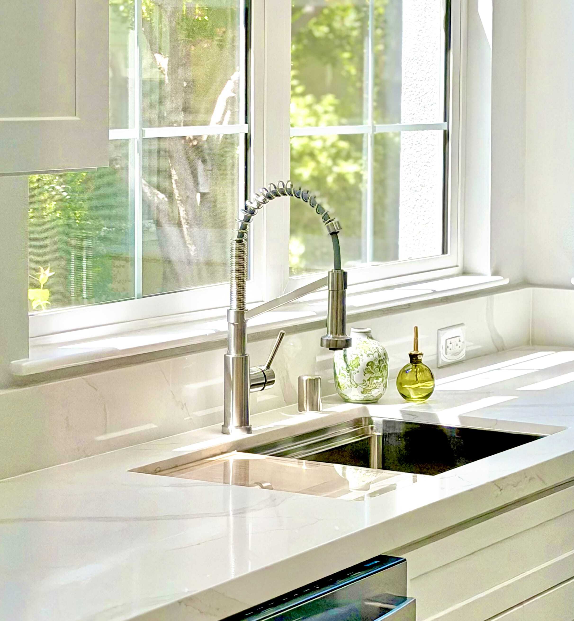 A modern kitchen sink with a tall, flexible faucet, set in a white marble countertop. Sunlight streams through a window above, and a green soap dispenser and plant sit on the counter.