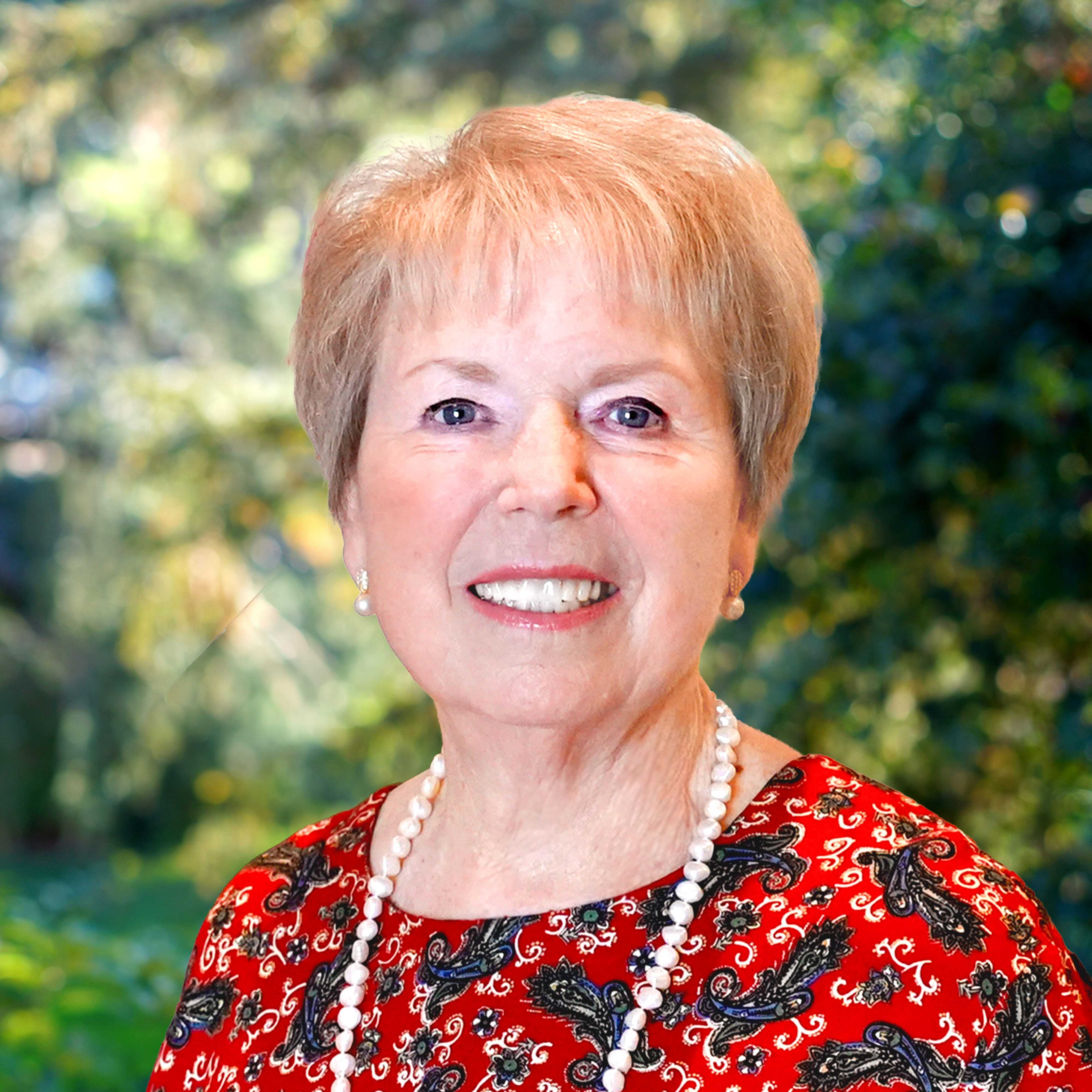 Smiling older woman with short light hair, wearing a red patterned top and pearl necklace, standing outdoors with greenery in the background.