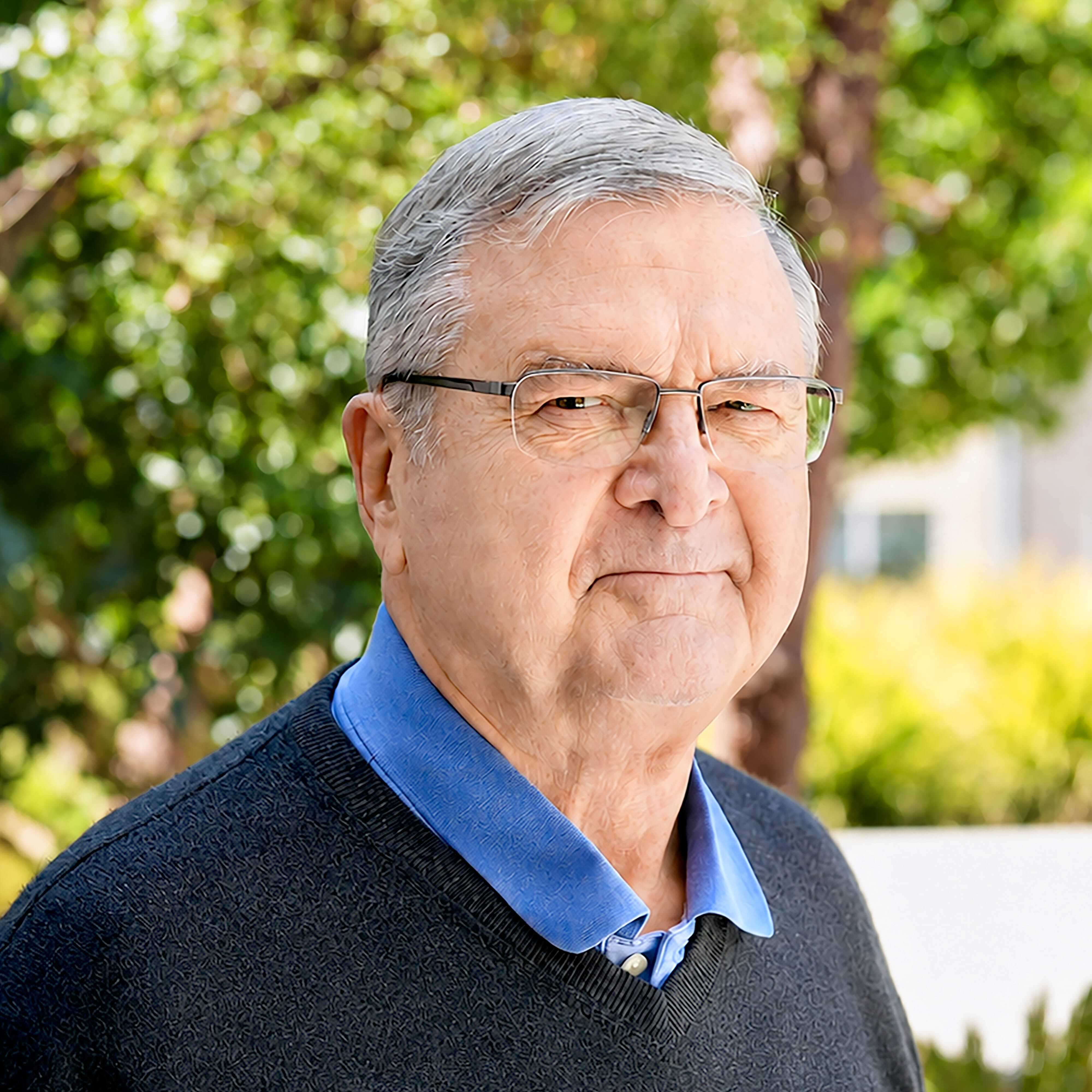 An older man with gray hair and glasses, wearing a blue collared shirt and a dark sweater, stands outdoors with trees and greenery blurred in the background.