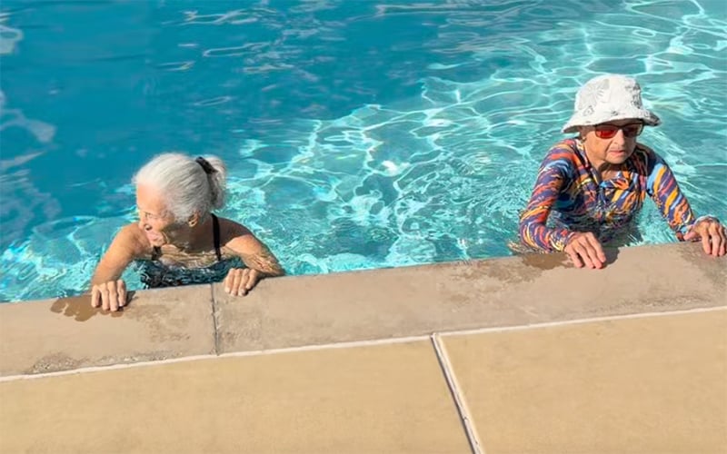 Screenshot Two elderly women hold onto the edge of a swimming pool. One wears a colorful shirt, white hat, and sunglasses; the other has gray hair tied back and wears a black swimsuit. Both are in clear blue water near the poolside.