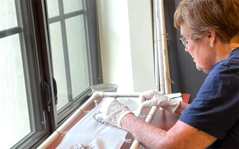 Screenshot An older person wearing glasses and gloves works on a craft project by a window, tracing or painting a design onto a white fabric or paper held in place by a PVC frame.