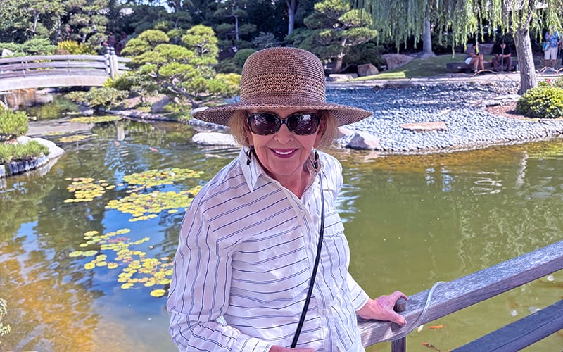 01-CAN-Header_Oct2026 A woman in a striped shirt, sunglasses, and straw hat smiles by a pond with lily pads in a sunny garden. There is a wooden bridge and lush greenery in the background.