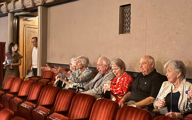 01-MC-Header_Oct2026 A group of six older adults sit in red theater seats, engaged in conversation. Two people stand near the entrance at the back. The mood appears casual and relaxed in a warmly lit auditorium.