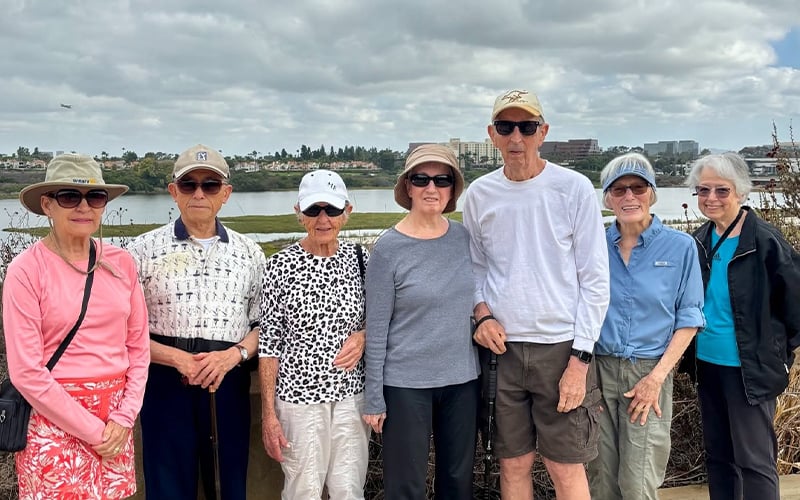01_COV-Header_Oct2026 Seven older adults in casual clothing and hats stand outdoors in front of a body of water, smiling at the camera on a cloudy day. Buildings and greenery are visible in the background.
