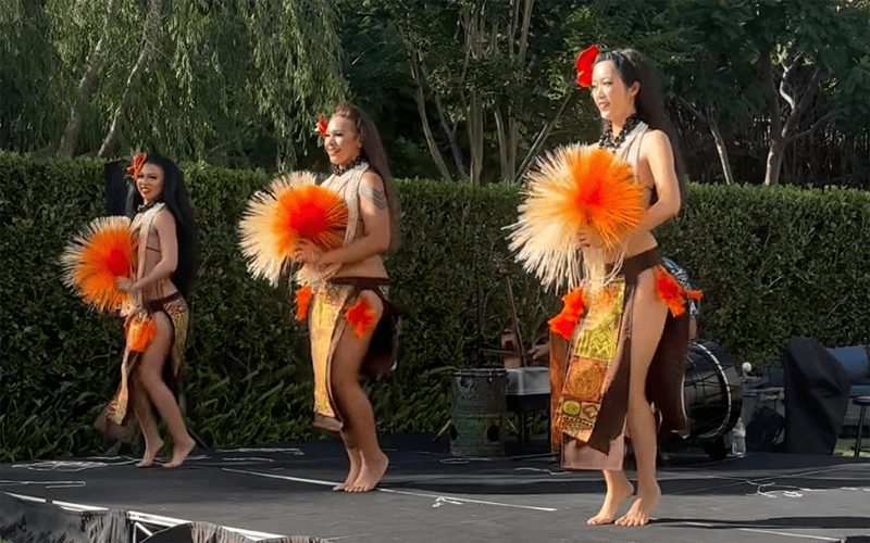 03-Story_01_COV_Luau Three women in traditional Polynesian attire perform a dance outdoors, holding orange and yellow feathered fans and wearing floral hairpieces. They are barefoot on a stage, with greenery in the background.