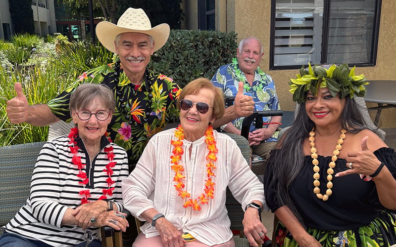03-Story_01_Luau 1 Five smiling seniors wearing Hawaiian shirts and leis pose outdoors. One man wears a cowboy hat, and one woman wears a leafy headpiece. They all give a thumbs-up gesture, enjoying a festive tropical-themed gathering.