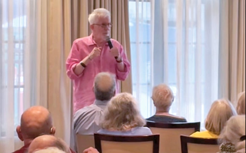 Screenshot A man in a pink shirt stands and speaks into a microphone in front of an audience of older adults seated indoors, near large windows with white curtains.