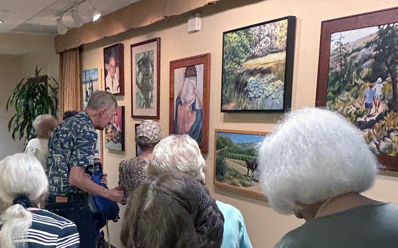 03-Story_02_COV_Sept Artist in Res A group of elderly people closely view various framed paintings displayed on a light-colored gallery wall. The art features landscapes and portraits.