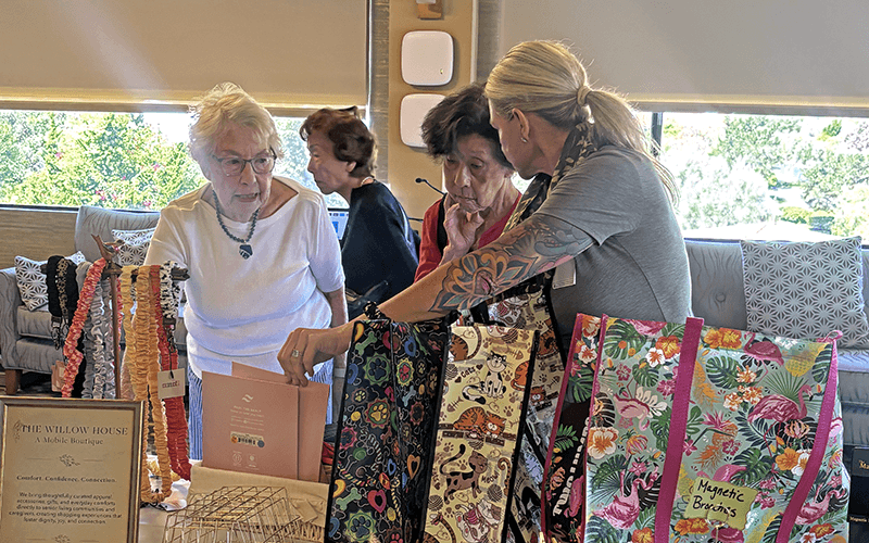 03-Story_02_WillowBoutique 2 Three women examine colorful fabric bags displayed by a vendor with tattooed arms at an indoor craft market. A sign on the table reads The Willow House. Sunlight filters through large windows in the background.