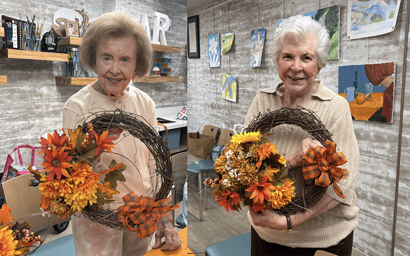 03-Story_03_GrapevineWreaths Two elderly women smile while holding autumn wreaths decorated with orange flowers and plaid bows in a craft room with art displayed on the wall behind them.