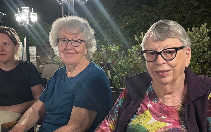 03-Story_04_COV_NeilDiamondConcert Three older women sit outdoors at night, two smiling at the camera and one looking away. They are surrounded by greenery and bright outdoor lights.