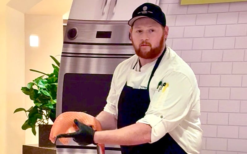 Screenshot A chef in a white coat and black apron holds a large fillet of raw salmon. He wears a black cap and gloves, standing in a kitchen with white tiled walls and a stainless steel oven behind him.