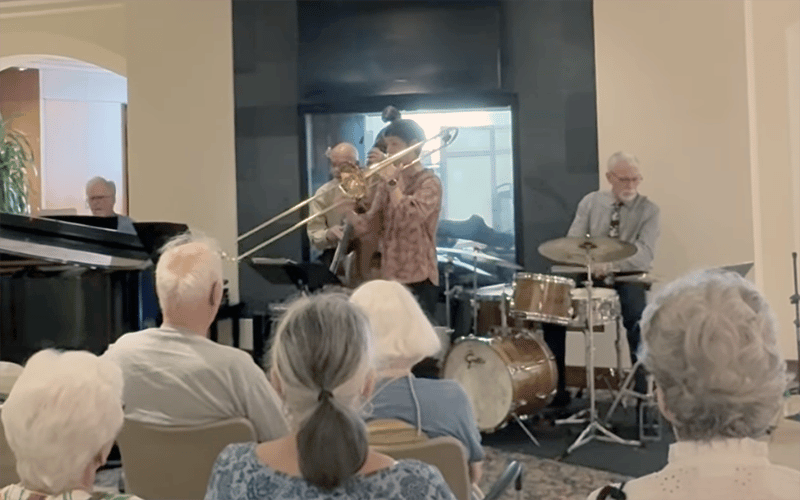03-Story_05-ChadE Jazz Band A small jazz band performs for an audience of seniors in a cozy indoor setting, with musicians playing trombone, piano, drums, and upright bass.
