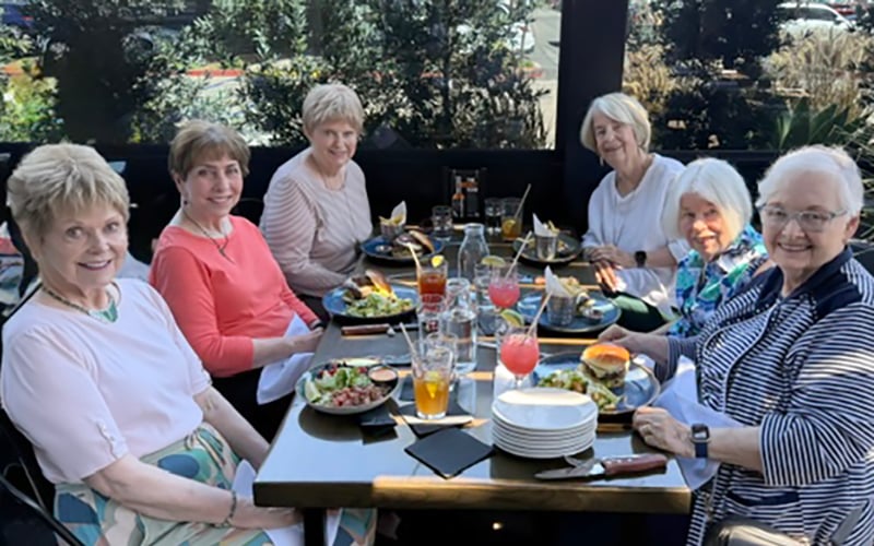 03-Story_06_Cov_Sept lunch in park Six older women sit around an outdoor restaurant table, smiling and enjoying a meal with salads, burgers, and colorful drinks. Sunlight filters through trees and plants in the background.