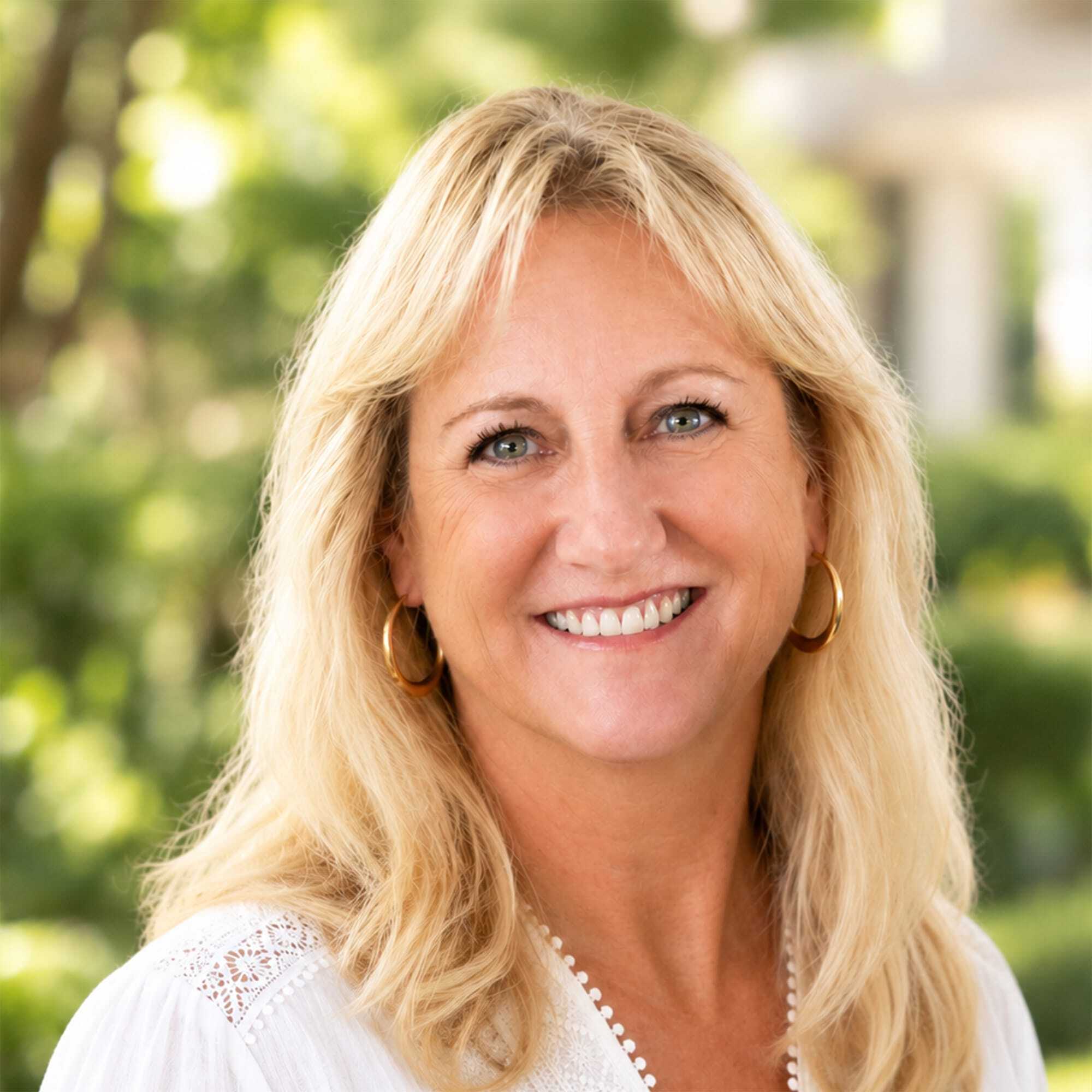 A smiling woman with long blonde hair wearing a white blouse and gold hoop earrings stands outdoors with blurred green foliage in the background.