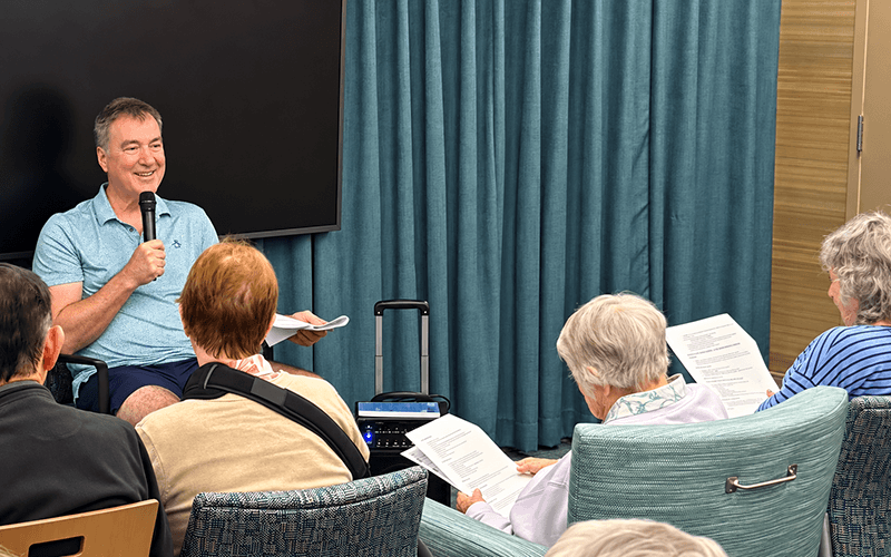 03-Story_01_CAN_TBurke Presentation_StoryTime A man speaks into a microphone in front of an audience holding papers in a conference room.