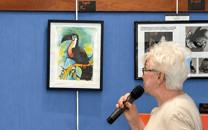 03-Story_02_CAN Artist Gallery_Toucan with Lynne A woman with short white hair holds a microphone while standing in front of a framed watercolor painting of a toucan.
