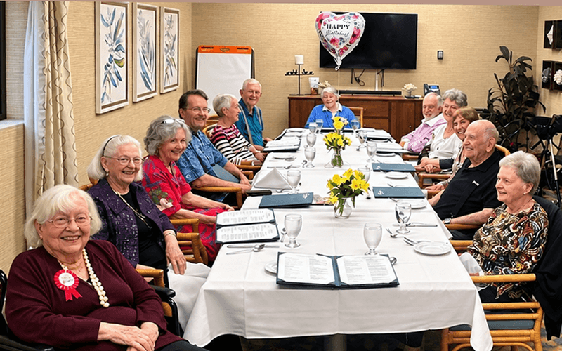 03-Story_03_CAN_Betty Birthday Luncheon A group of seniors sitting around a long, white-clothed table for a celebration with a Happy Birthday balloon.