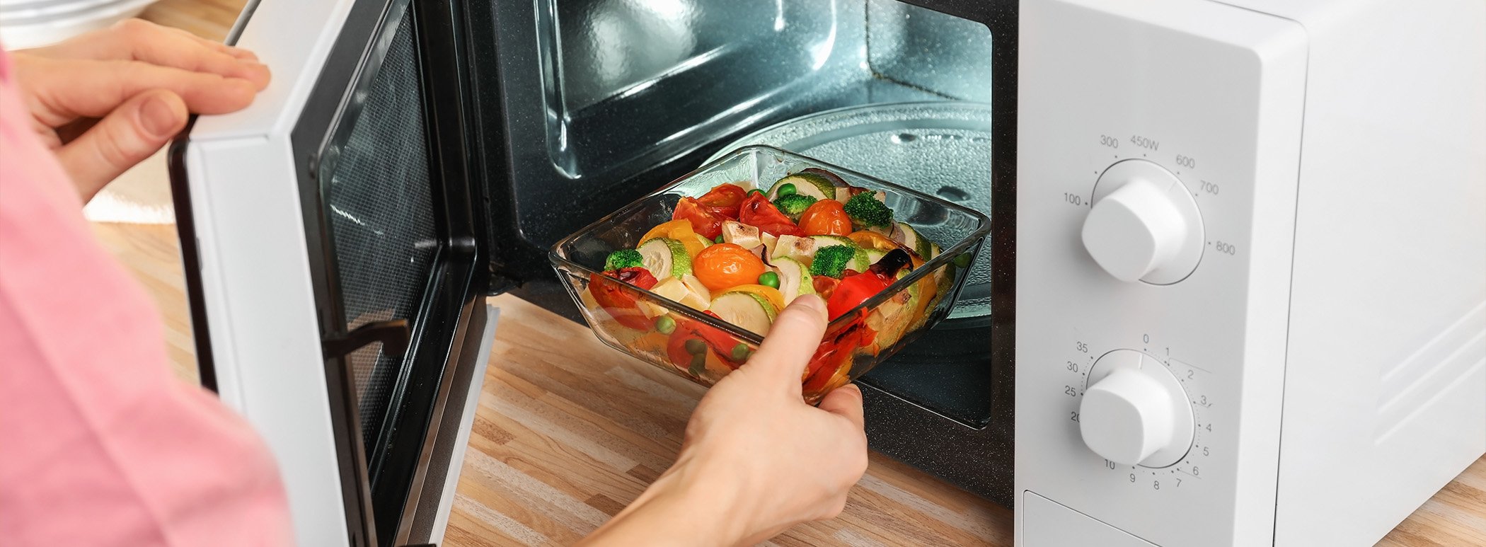A person places a glass dish of roasted vegetables into a microwave.