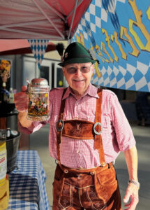 An older man wearing traditional Bavarian clothing, including lederhosen, a red checkered shirt, and a green hat with a feather, smiles while holding up a large beer stein under a red canopy decorated with blue and white Bavarian flags.