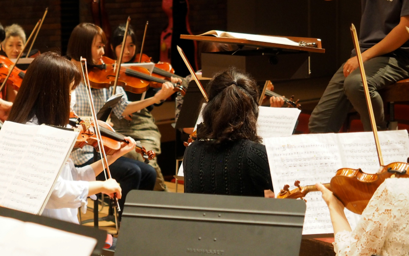 Several musicians, mostly women, are seated and playing violins in an orchestra rehearsal. Sheet music is visible on stands, and a conductor or instructor is partly visible at the front.