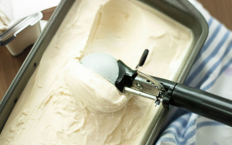 A metal ice cream scoop is being used to scoop vanilla ice cream from a rectangular metal container. The container is placed on a surface next to a striped cloth.