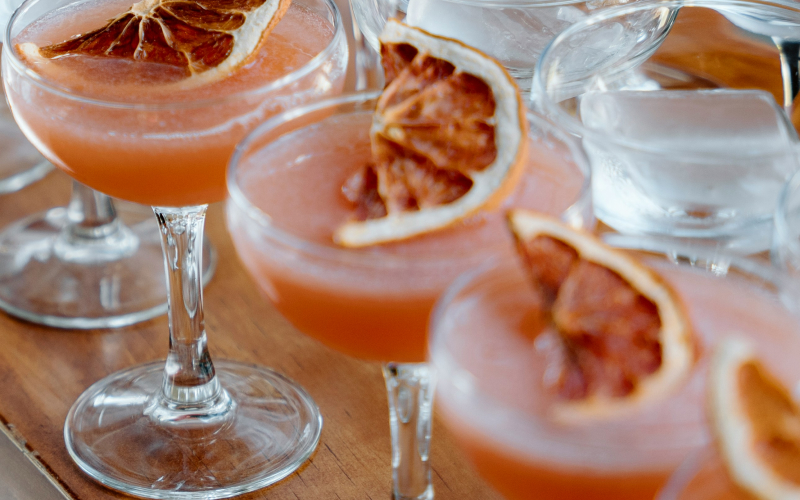 Cocktail glasses filled with a pink-orange drink, each garnished with a dried citrus slice, sit on a wooden surface surrounded by clear ice cubes.