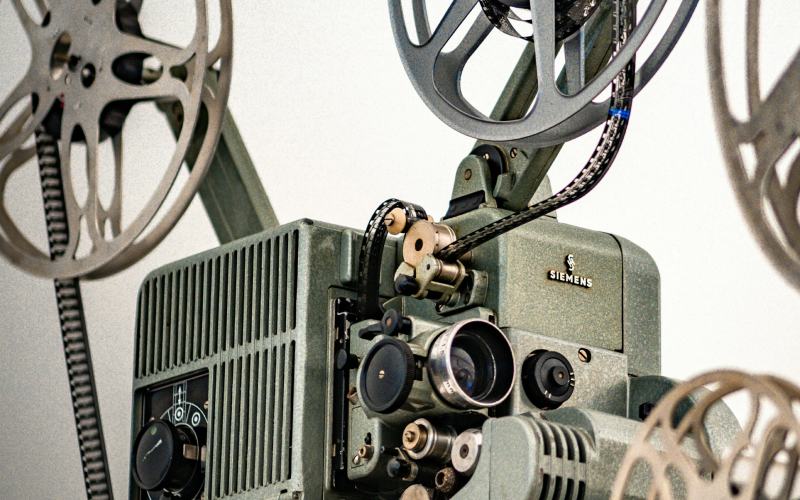 Close-up of a vintage Siemens film projector with metal reels, various knobs, dials, and a visible film strip threaded through the machine, set against a plain light background.