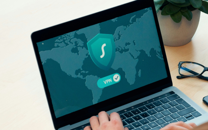 A person uses a laptop displaying a VPN shield logo and a world map on screen. Glasses and a plant are nearby on the desk.
