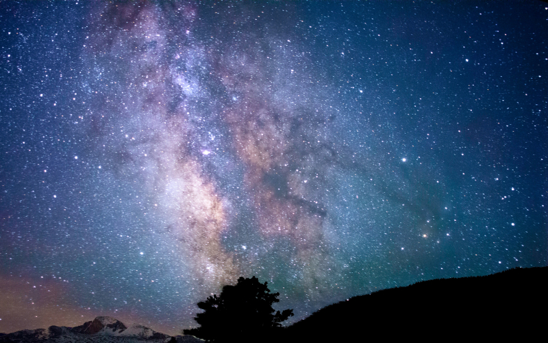 The night sky filled with stars and the vibrant colors of the Milky Way galaxy, with silhouetted mountains and a tree in the foreground.